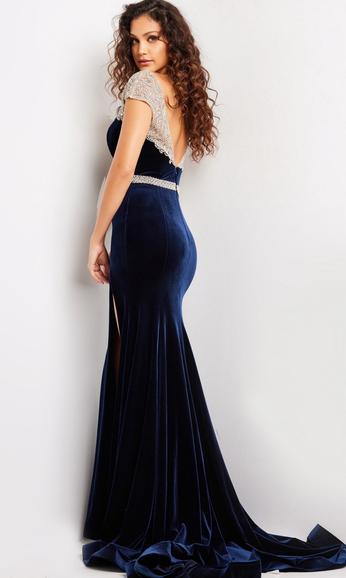A woman in the Long Prom Dress 37246 by Jovani, featuring navy blue crystal-embellished velvet, beaded cap sleeves, and an open back, stands against a light background, looking over her shoulder.
